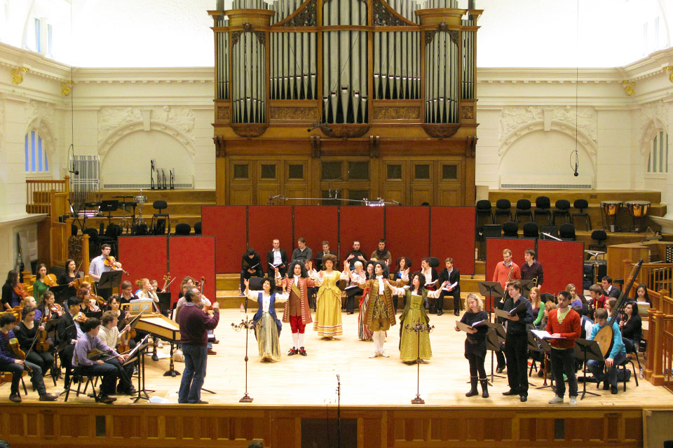Students rehearsing for a musical performance on either sides of the stage, with dancers dressed in period costume, with two singers performing at the top of the stage.